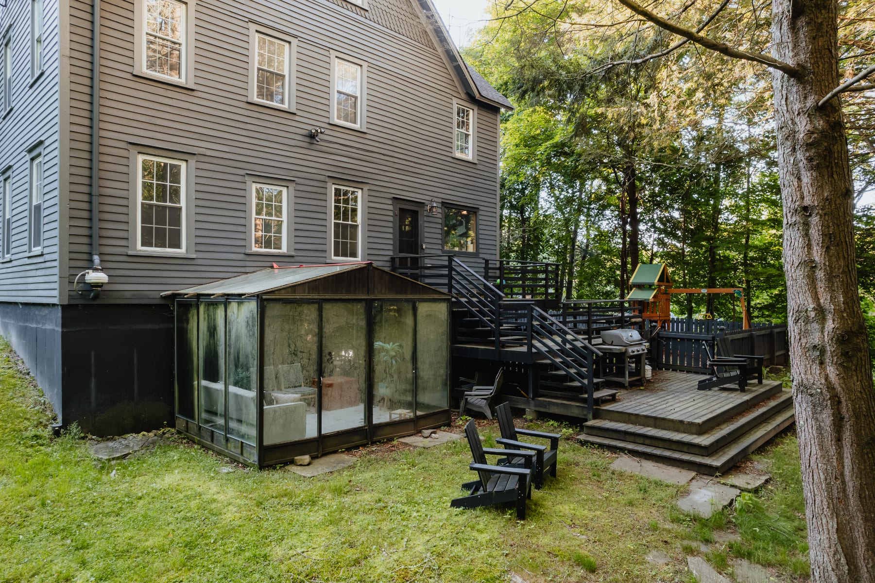A full view of the dark gray farmhouse's back exterior, showcasing a multi-level wooden deck with stairs, a BBQ grill, and a glass greenhouse, all surrounded by a lush green lawn and mature trees.