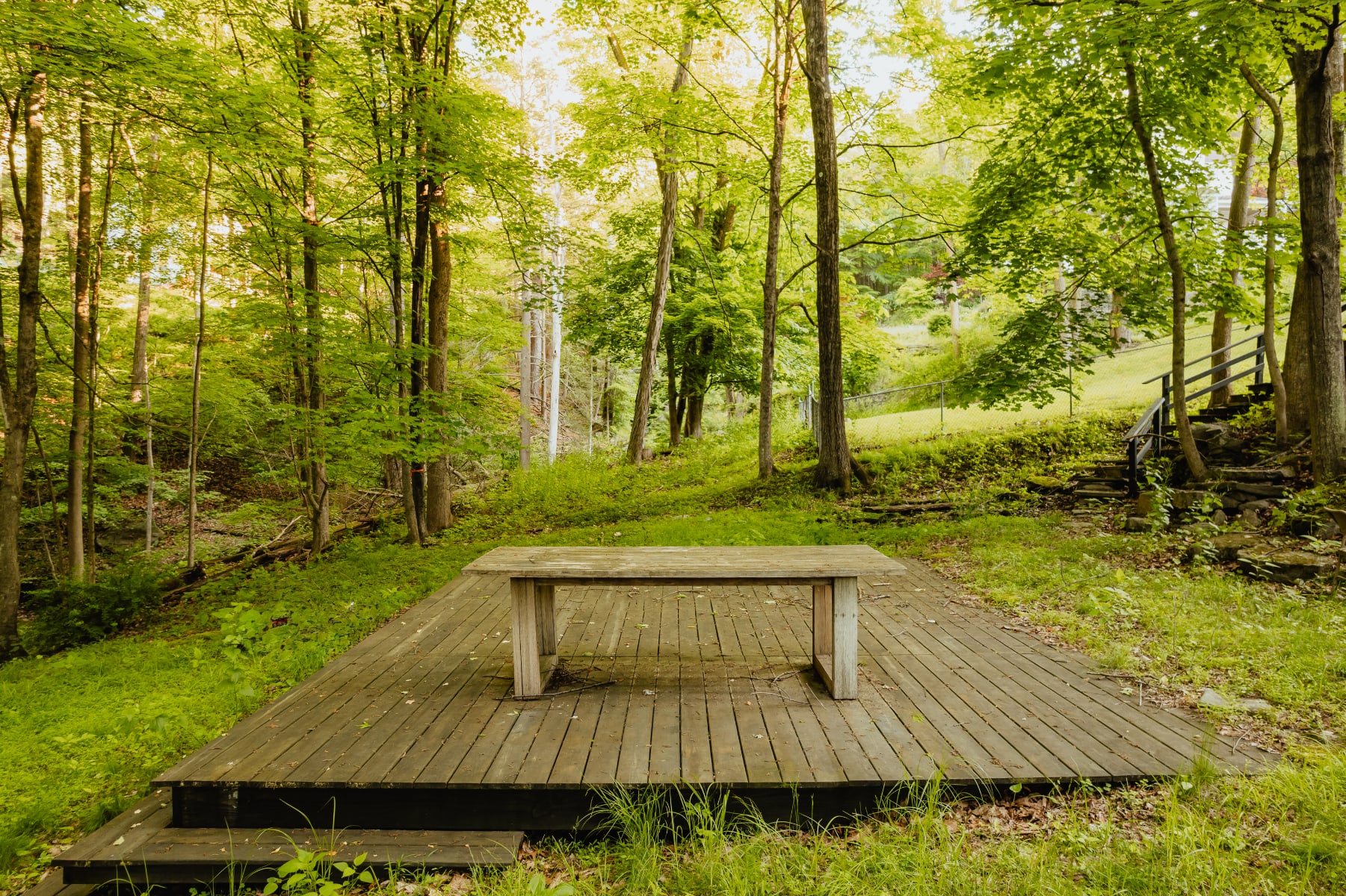 A large wooden deck platform, possibly for outdoor dining or activities, is nestled in a lush green, wooded area, featuring a long wooden table in the center.