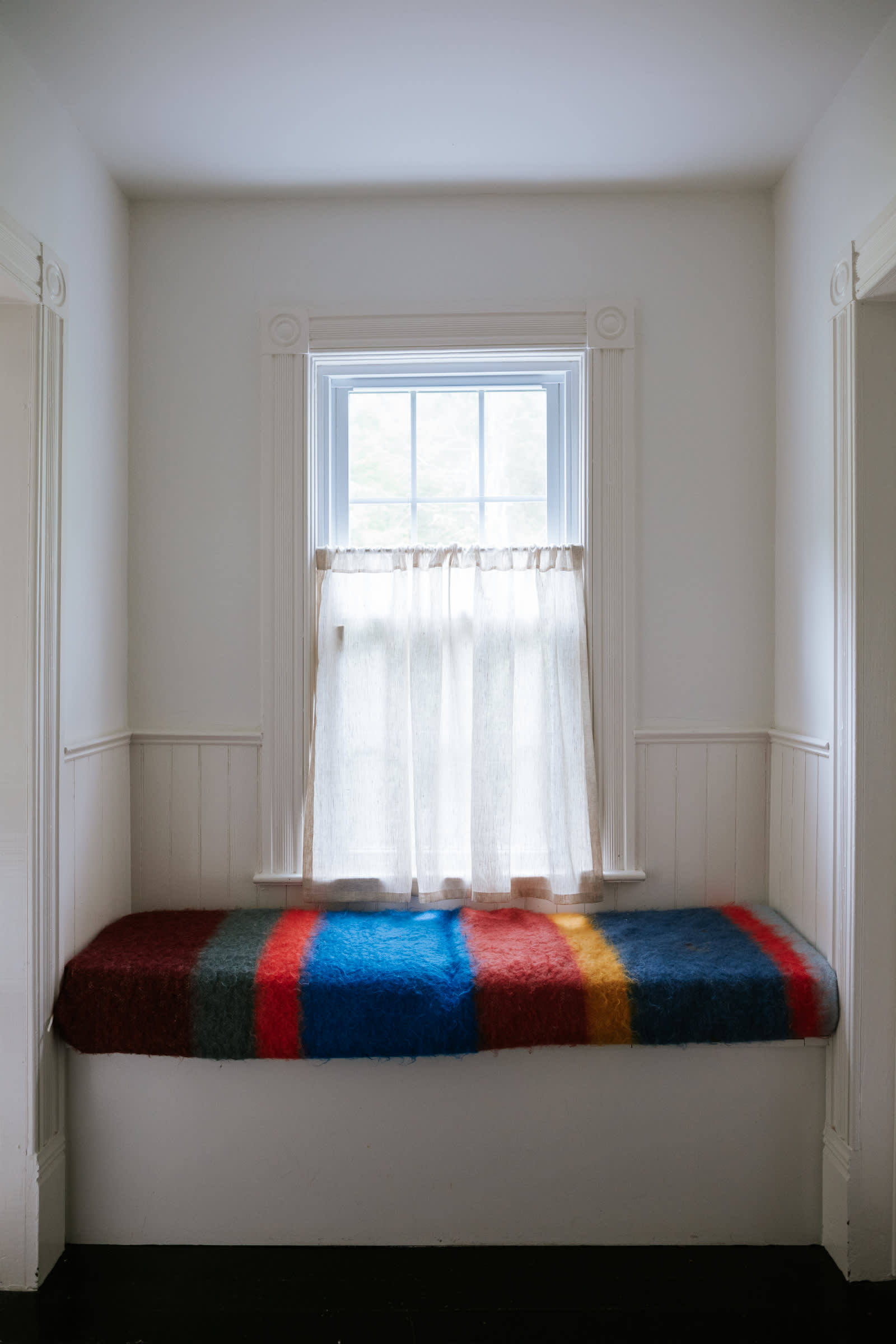 A bright alcove with white walls and wainscoting, featuring a built-in window seat covered with a vibrant striped blanket in red, blue, yellow, and green hues. A single window with sheer curtains provides natural light.