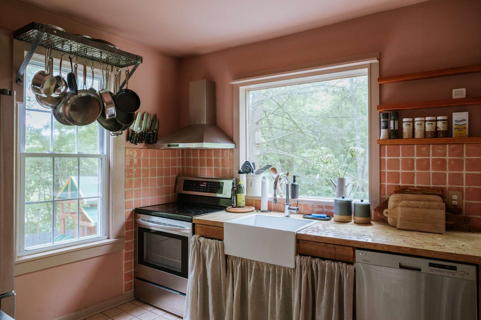 A rustic kitchen with pink walls and terracotta-colored square tile backsplash features a white farmhouse sink, stainless steel appliances, and wooden countertops. Pots and pans hang from a rack above the stove.