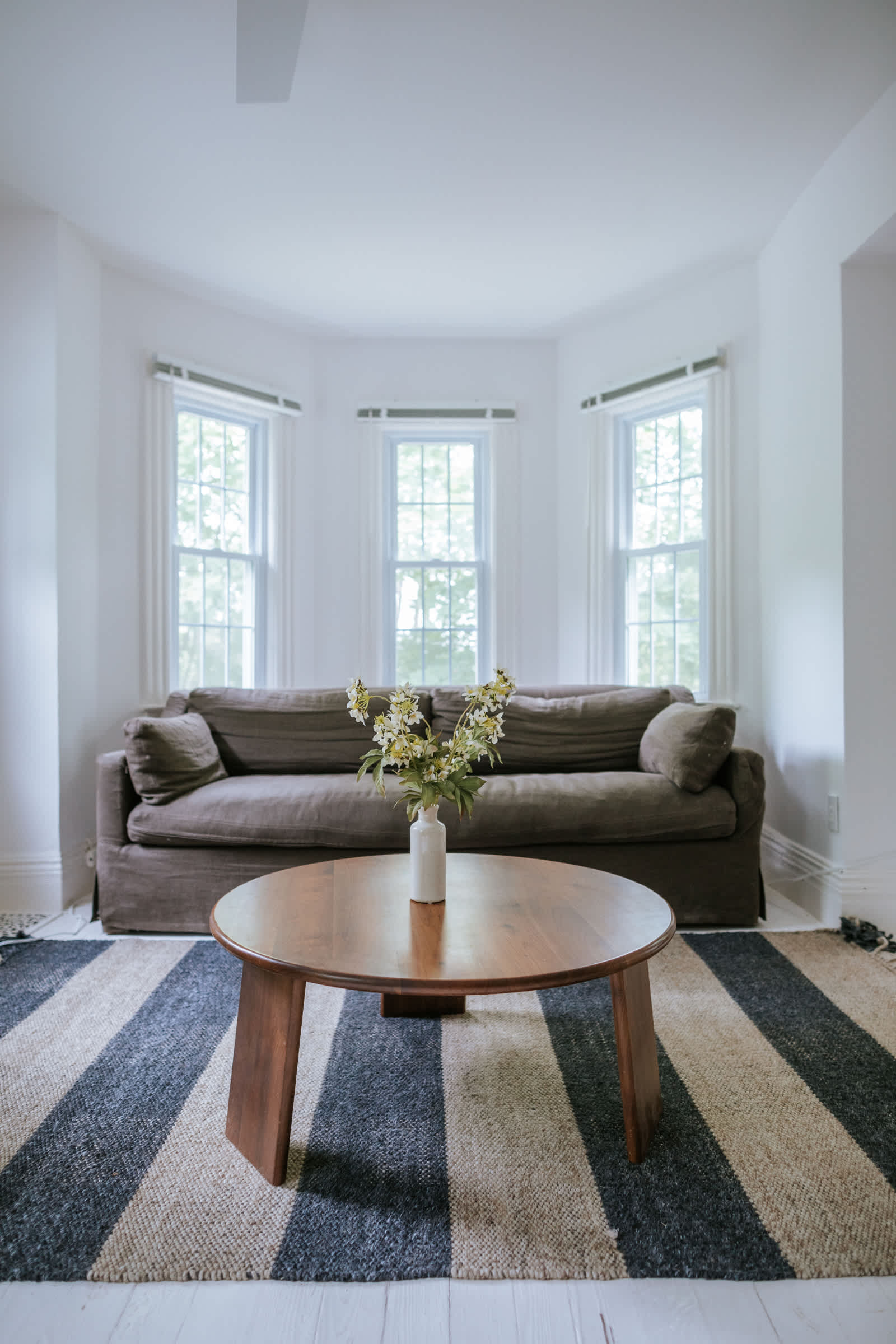A cozy sitting area within a bedroom, featuring a dark grey slipcovered sofa nestled in a bay window alcove with three large windows. A round wooden coffee table with a white vase of flowers sits on a striped black and natural fiber rug.