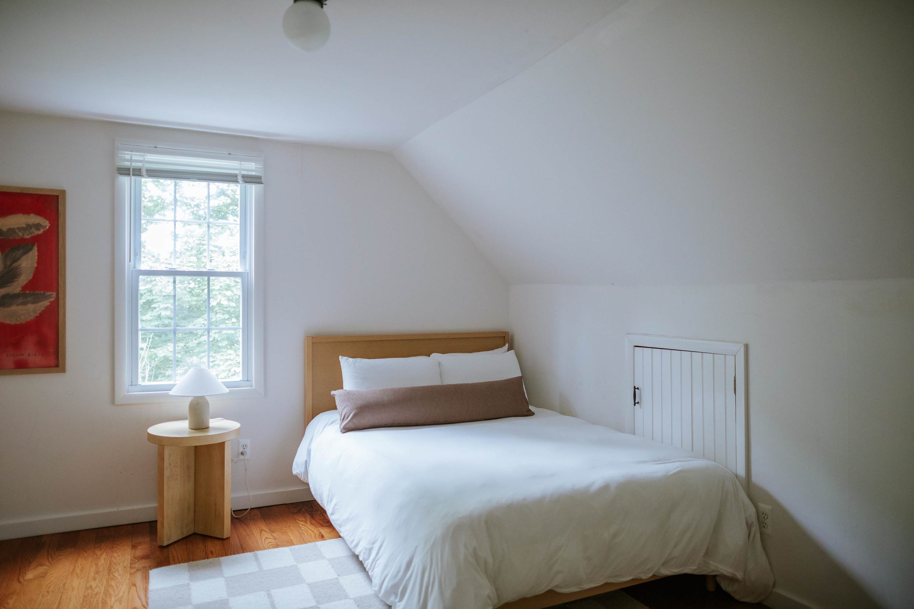 A cozy bedroom with white walls and a sloped ceiling features a light wood bed frame with a beige headboard, white bedding, a light wood round side table, and a white table lamp next to a window with blinds.