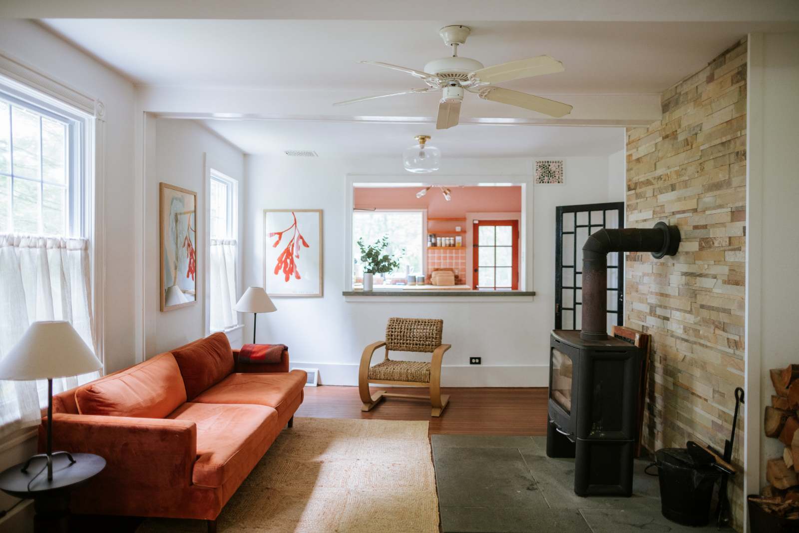 A cozy living room features an orange velvet sofa, a black wood-burning stove with a stacked stone-tiled wall, and a pass-through window to a pink-walled kitchen. Hardwood floors are partially covered by a natural fiber rug.