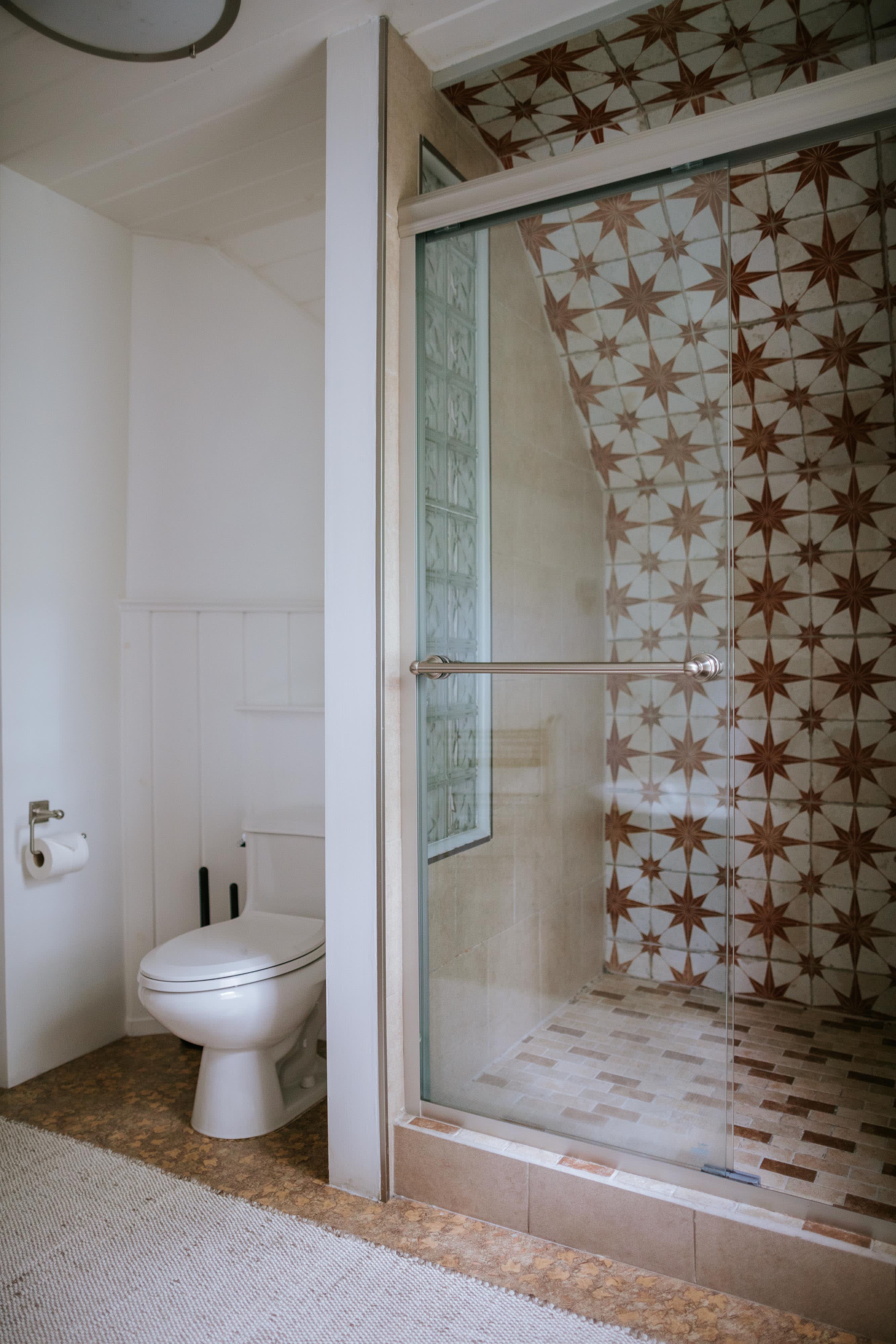 A bright bathroom features a walk-in shower with glass doors and distinctive star-patterned brown and beige tiles, a white toilet, and a glass block window. The floor is cork-like.