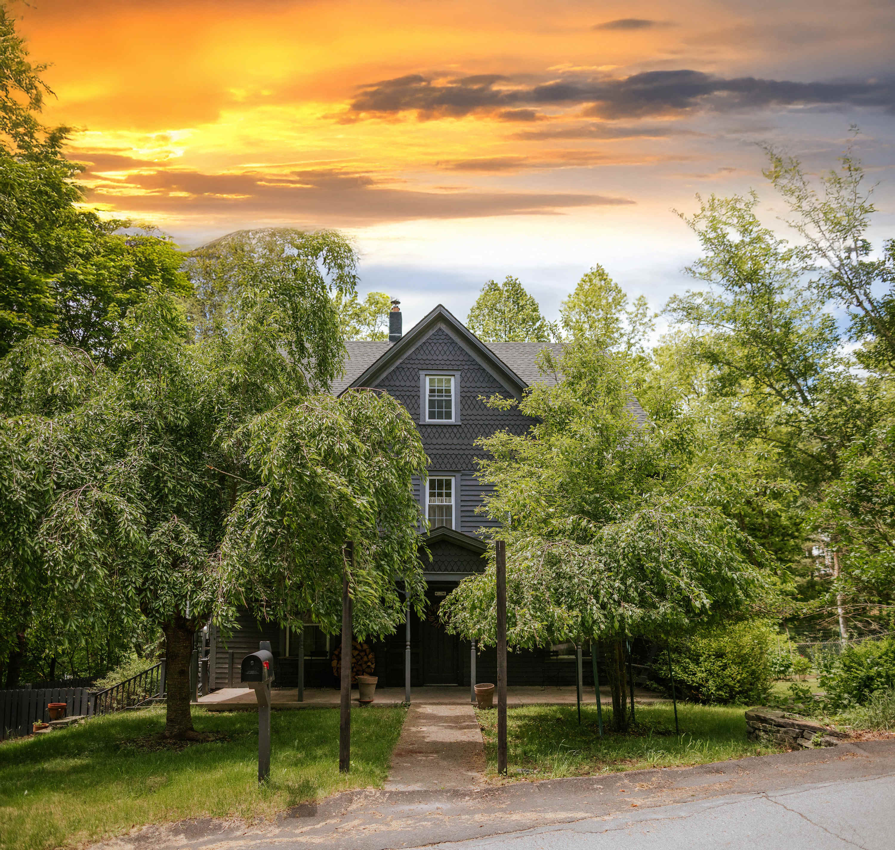 The front of the dark blue farmhouse is framed by lush green trees, with a concrete pathway leading to the entrance. A mailbox stands near the street, and a vibrant sunset sky is visible above the roofline.