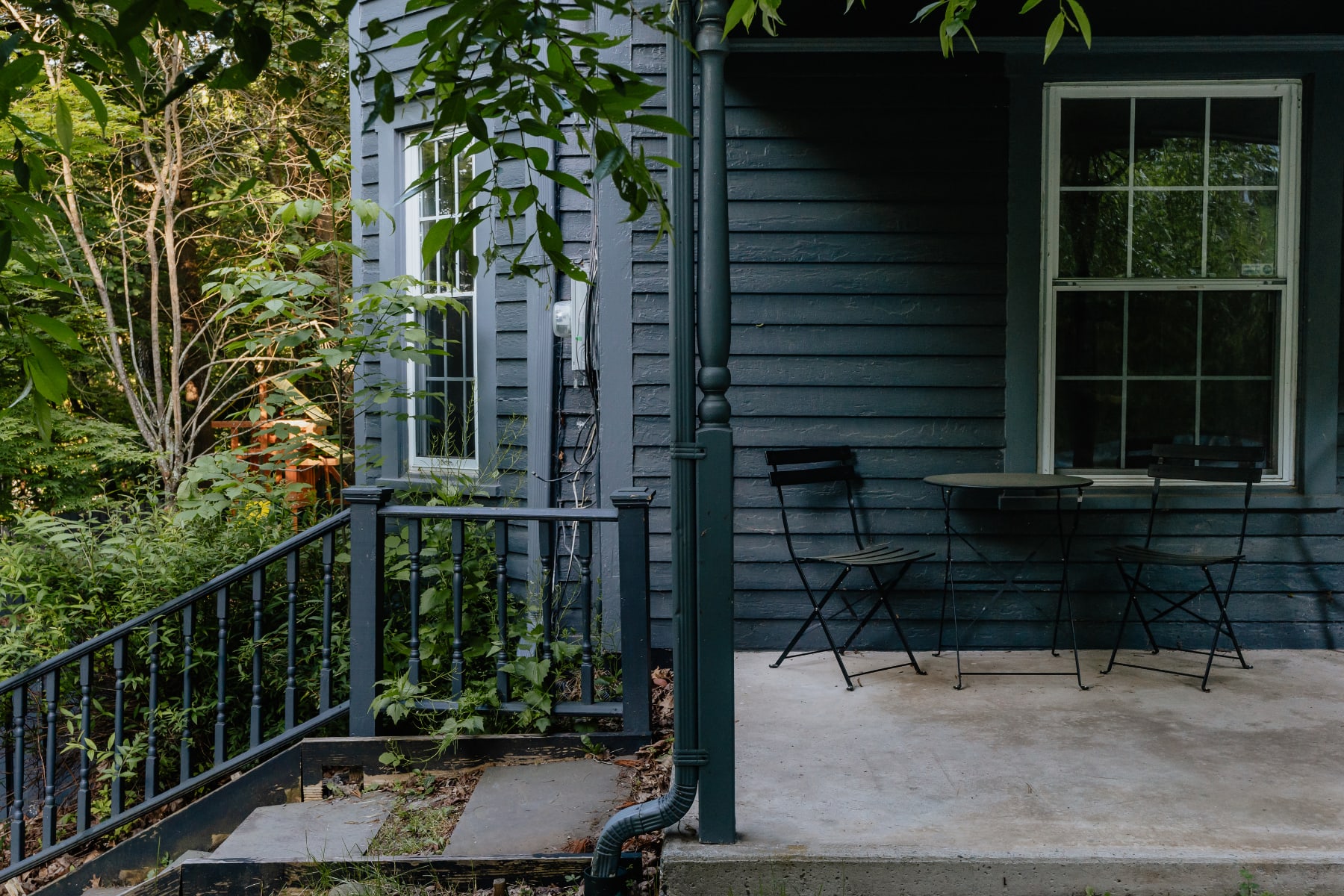 A dark gray farmhouse porch features a small bistro table with two chairs, set against the house's dark siding and white-trimmed windows, surrounded by lush green foliage.