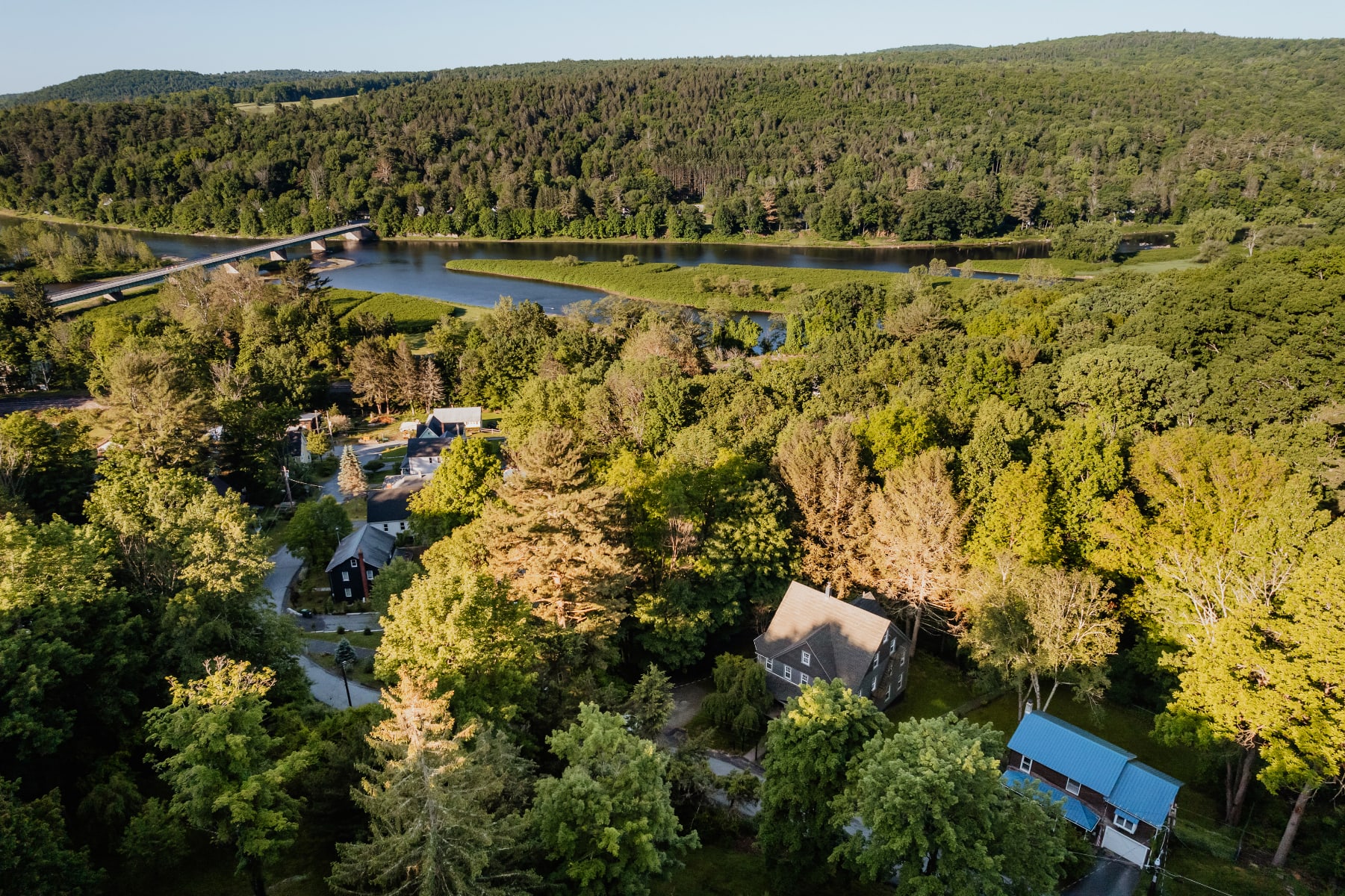 An aerial view captures the dark gray farmhouse nestled among dense green trees, with a river and bridge visible in the distance, highlighting the property's secluded, natural setting and water views.
