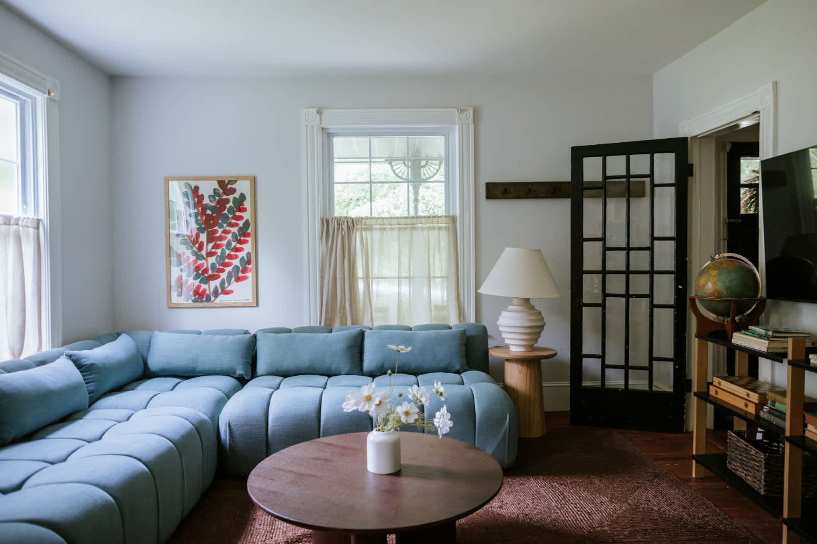 A bright living room features a large light blue sectional sofa, a round wooden coffee table with a vase of flowers, and two windows with sheer curtains. A black decorative door and abstract artwork adorn the white walls.