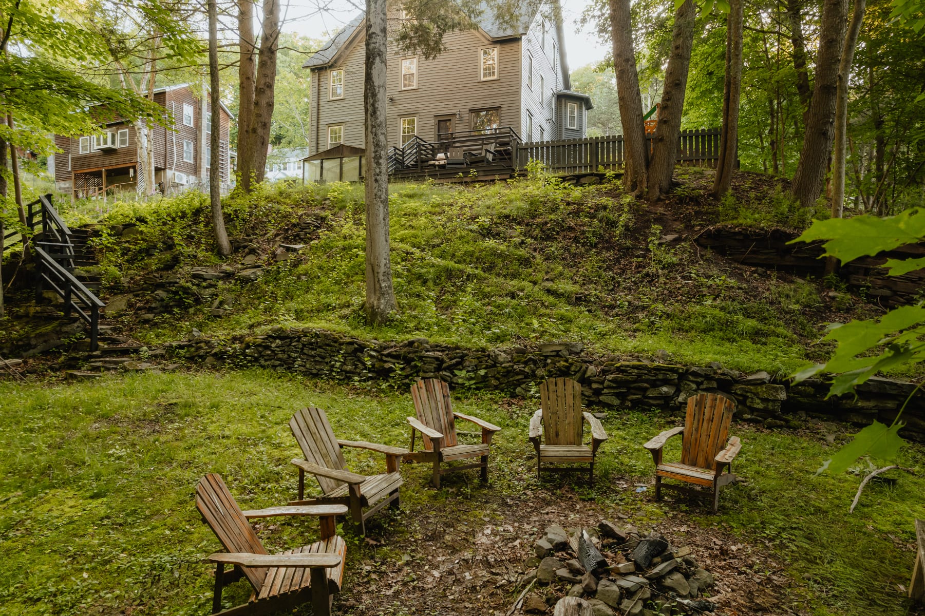 A rustic fire pit area in a grassy, wooded backyard features five wooden Adirondack chairs arranged around a stone fire pit, with the dark gray farmhouse visible on a hill in the background.