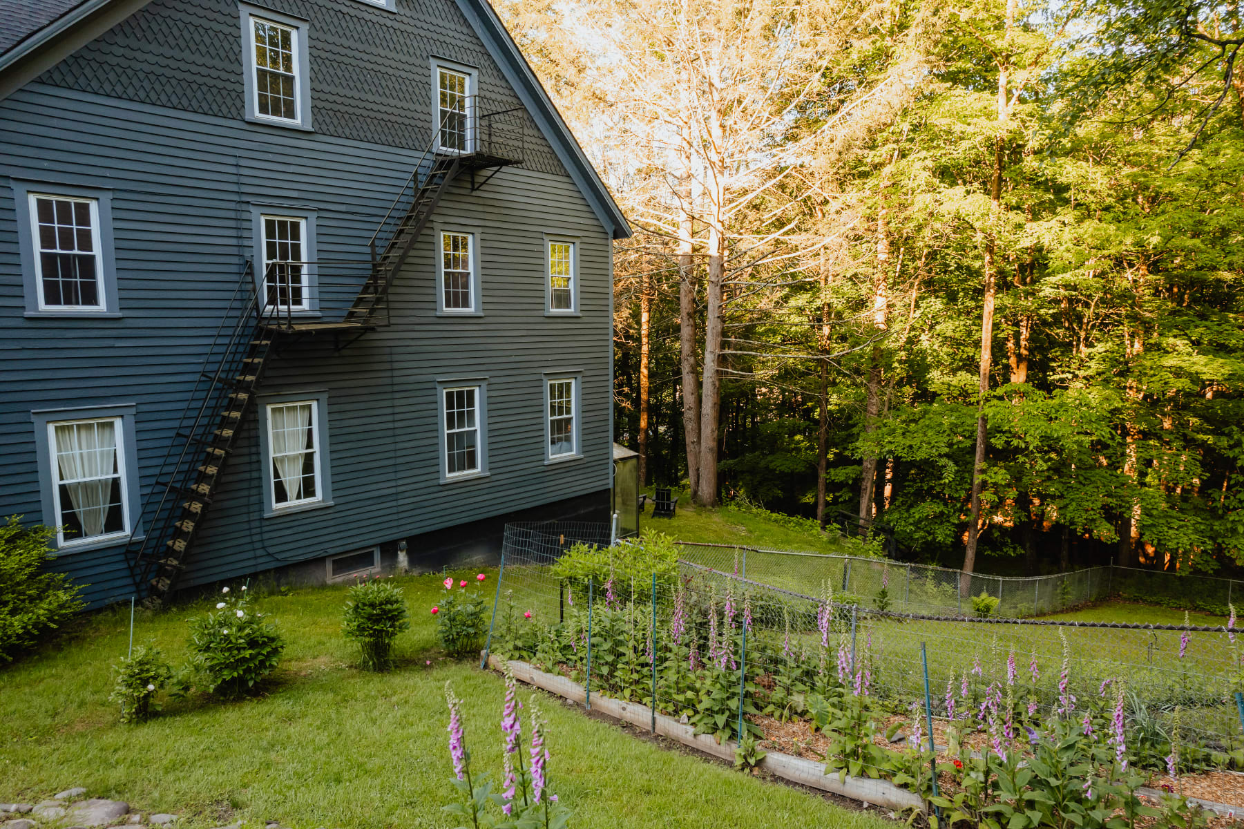 The side exterior of the dark gray farmhouse features a fire escape staircase and a vibrant garden bed with purple flowers and green plants, enclosed by a chain-link fence, leading to a wooded area.