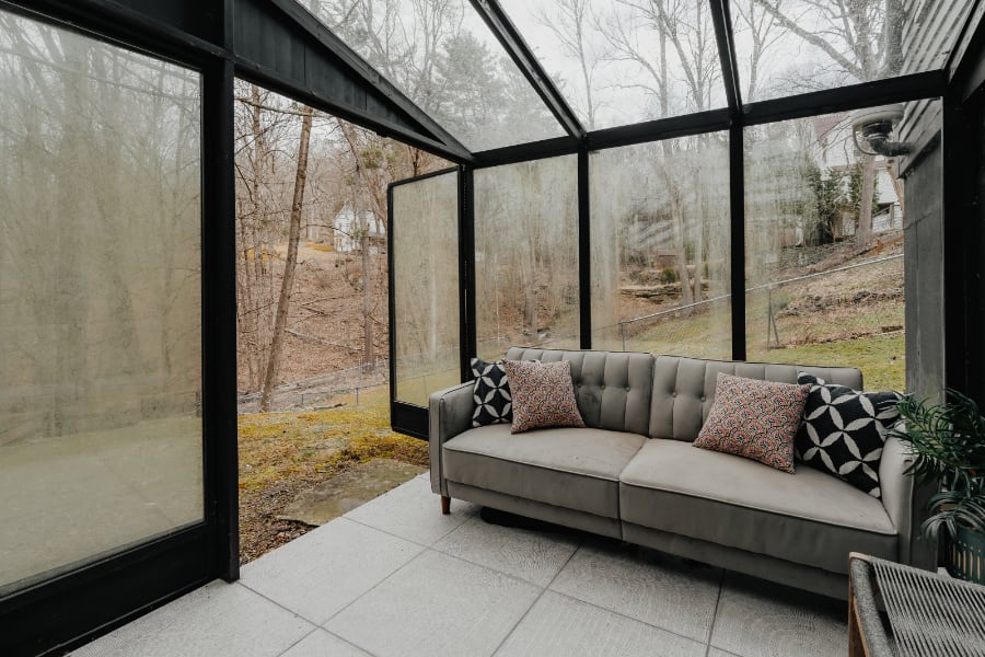 Sunroom with a gray tufted sofa adorned with patterned throw pillows, set against large glass walls and ceiling offering views of a wooded, overcast landscape. The floor is covered with white tiles.