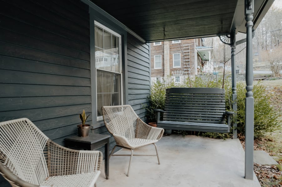 A cozy covered porch of a dark gray farmhouse features two woven armchairs and a small side table with a plant, alongside a dark wooden porch swing, all set on a concrete floor.