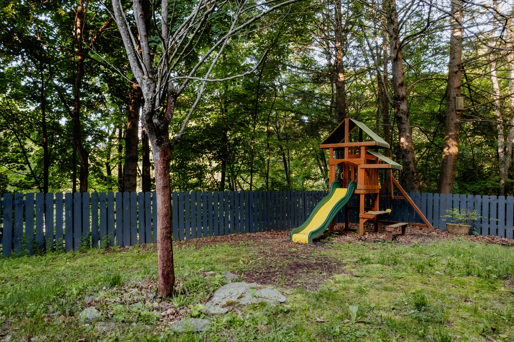 A children's wooden playset with a yellow slide is situated in a grassy backyard, enclosed by a dark gray picket fence and surrounded by tall trees, offering a family-friendly outdoor space.