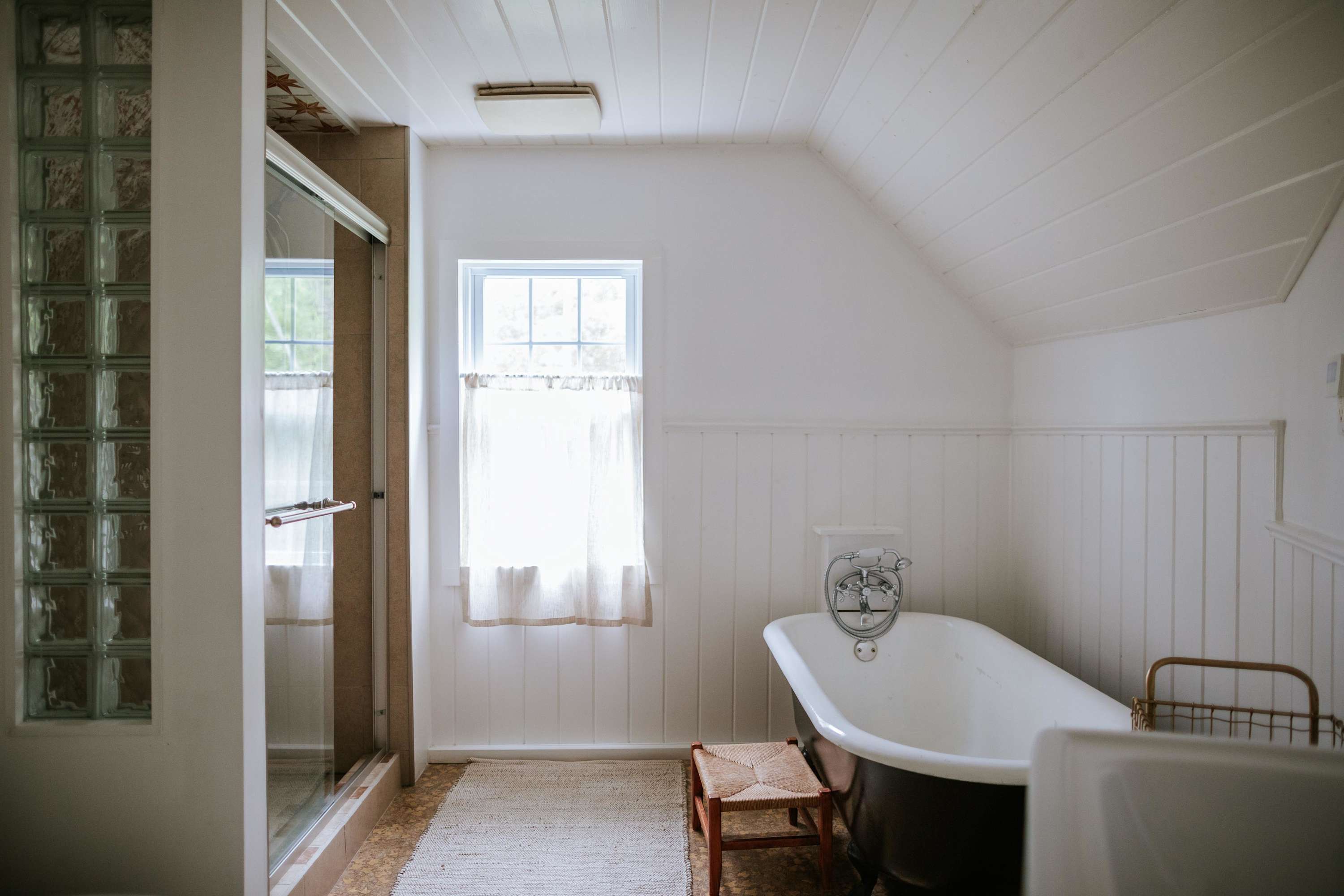 A bright bathroom with white paneled walls and a sloped ceiling features a classic black clawfoot bathtub, a walk-in shower with glass doors, and a window with sheer curtains.