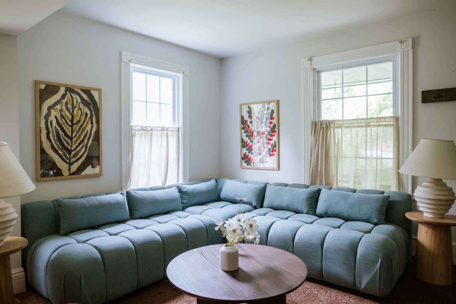 A bright living room features a large light blue sectional sofa, a round wooden coffee table with a vase of flowers, and two windows with sheer curtains. Abstract artwork hangs on the white walls.