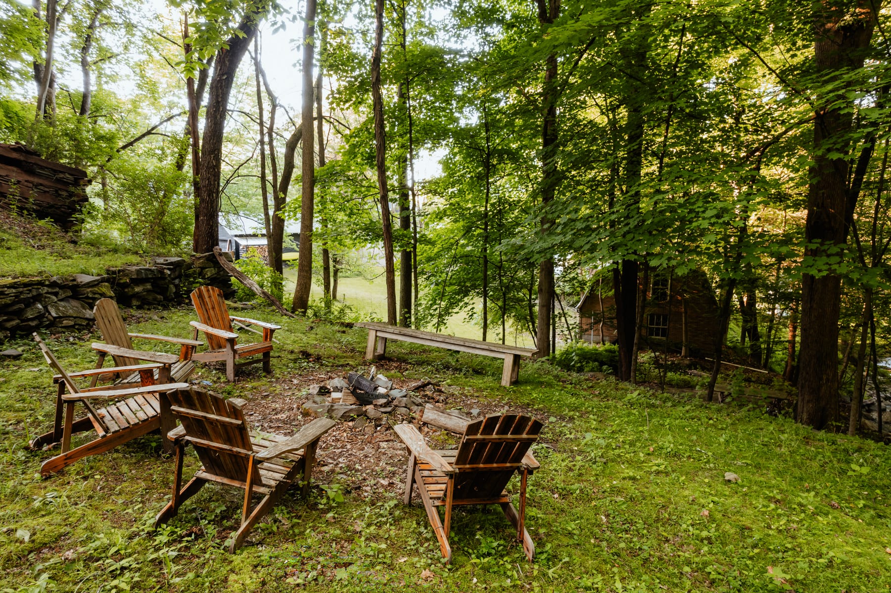 Outdoor fire pit area surrounded by lush green grass and tall trees, featuring several wooden Adirondack chairs and a long wooden bench. A rustic cabin is partially visible in the background through the foliage.