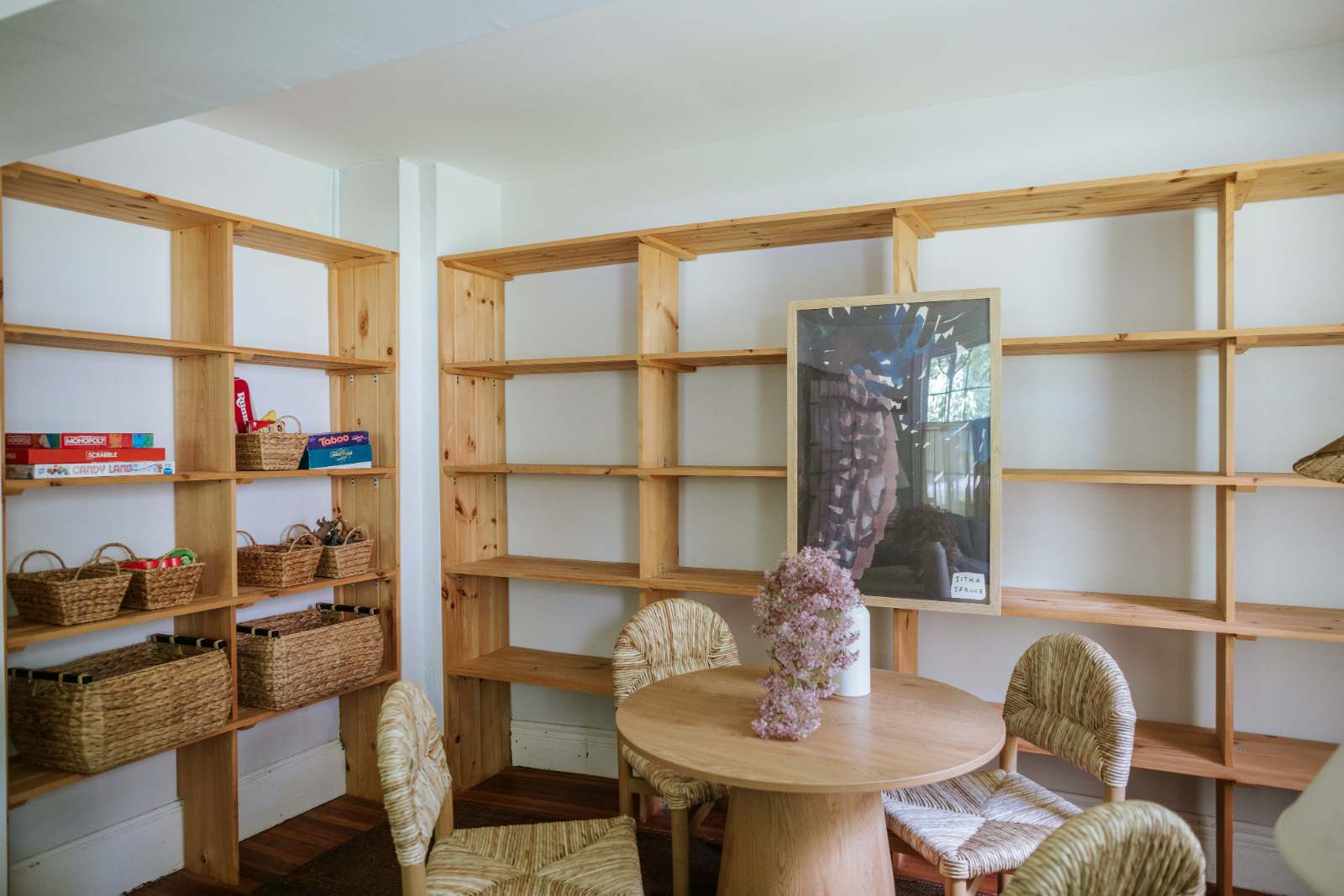 Bright room featuring extensive light wooden shelving filled with board games and woven baskets, a round wooden table, and four chairs with woven seats. A framed picture and vase of flowers decorate the table.