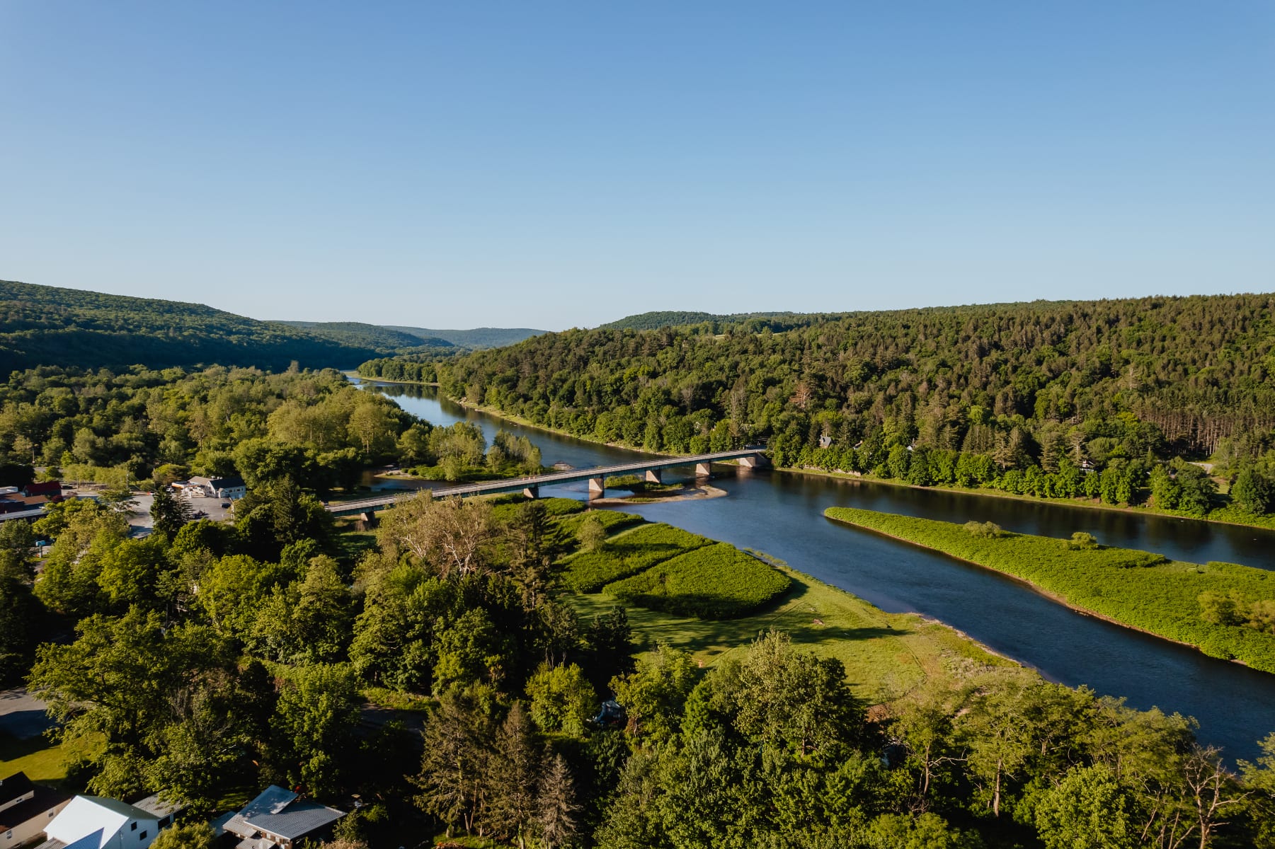 A stunning aerial view showcases a wide river with a bridge spanning across it, surrounded by dense green forests and a small town. The clear blue sky highlights the natural beauty of the landscape.