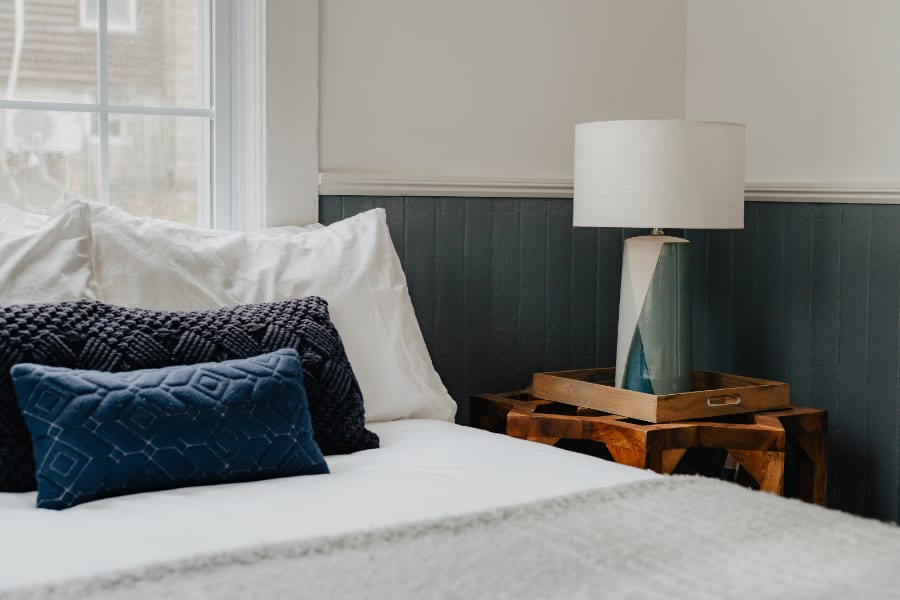 Close-up of a bedside area in a bedroom, showing a wooden side table with a decorative tray, a modern lamp with a white shade and blue base, and a bed with white and blue bedding.