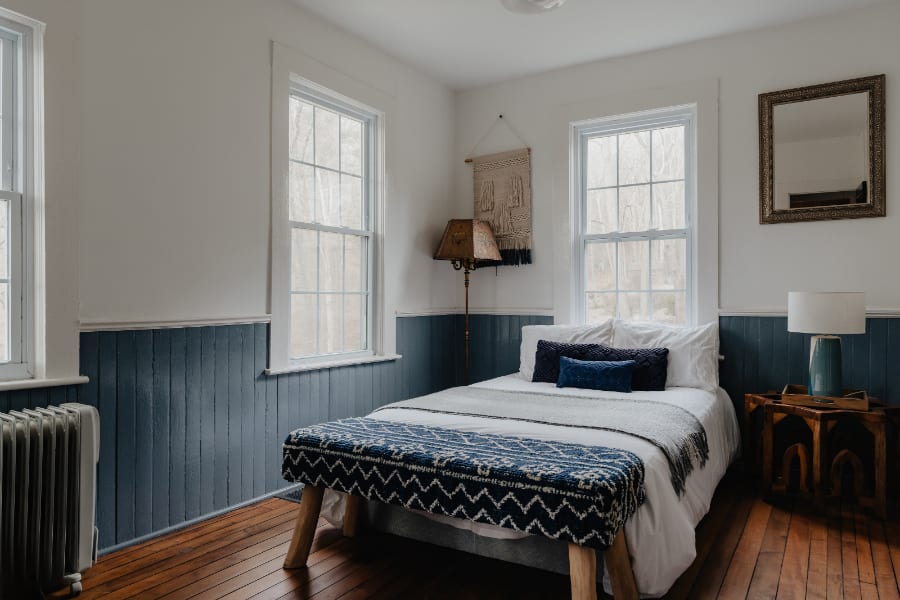 Bright bedroom featuring a bed with white bedding and blue accent pillows, dark blue wainscoting, and white walls. Two large windows overlook a wooded area, and a vintage-style floor lamp stands in the corner.
