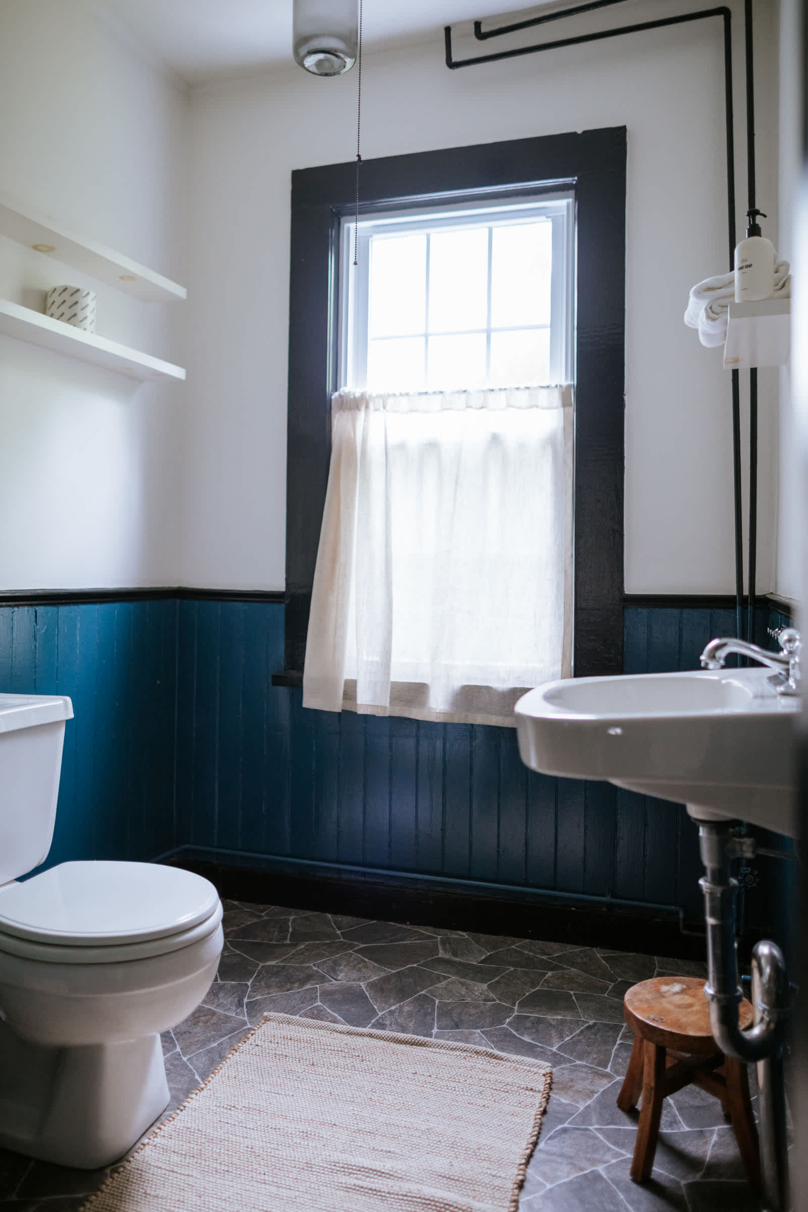 A compact bathroom features a white toilet, a pedestal sink, and a window with a dark frame and sheer curtain, set against white walls with dark blue wainscoting.