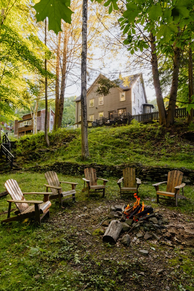 A cozy fire pit with burning flames is surrounded by five wooden Adirondack chairs in a grassy, wooded area, with the dark gray farmhouse visible on a hill in the background under warm sunlight.