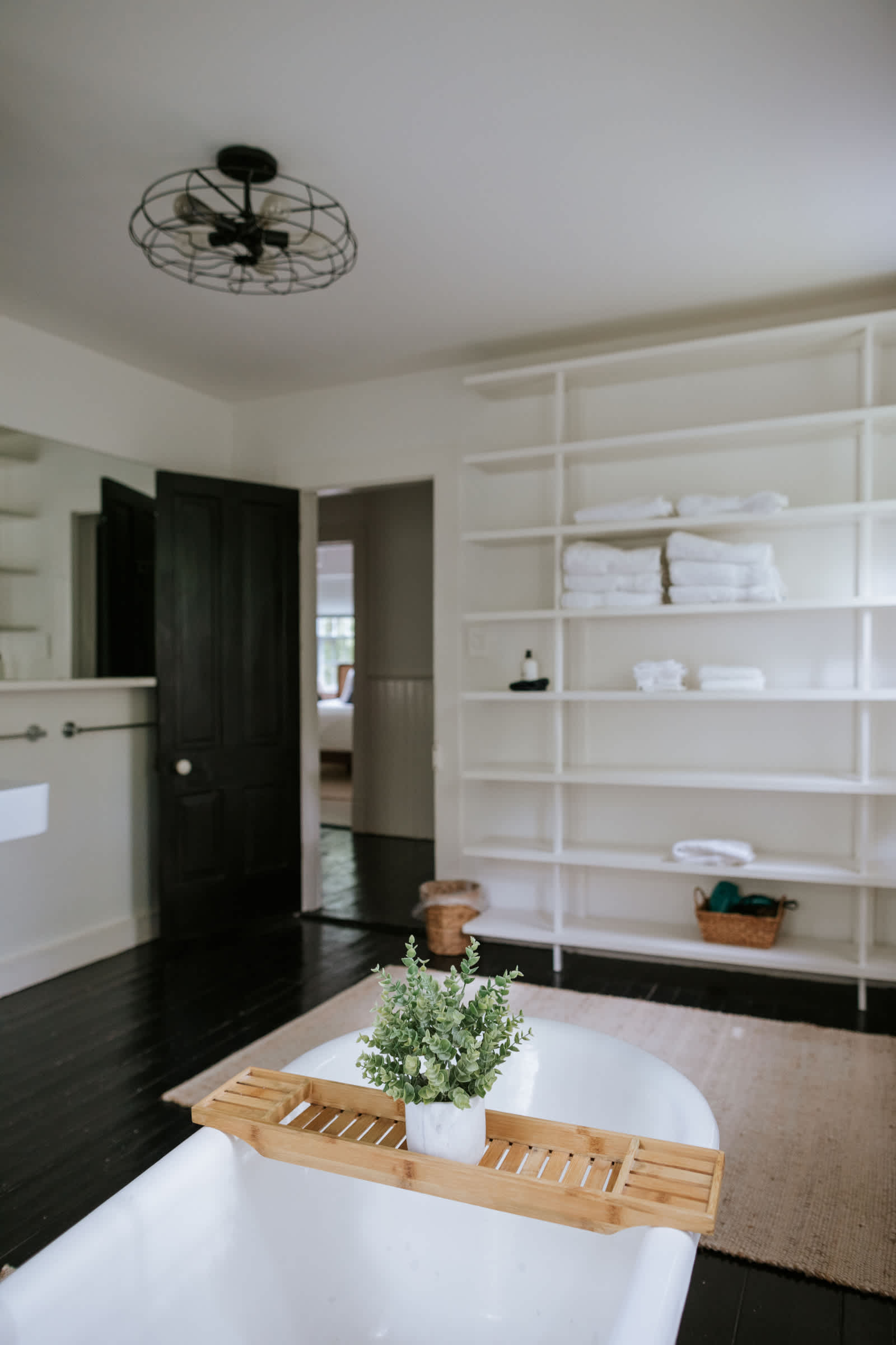 A wide view of a bright bathroom with black hardwood floors, featuring a white clawfoot tub in the foreground and a large white built-in shelving unit filled with neatly folded towels and toiletries. A dark door leads to another room, and a ceiling fan is overhead.