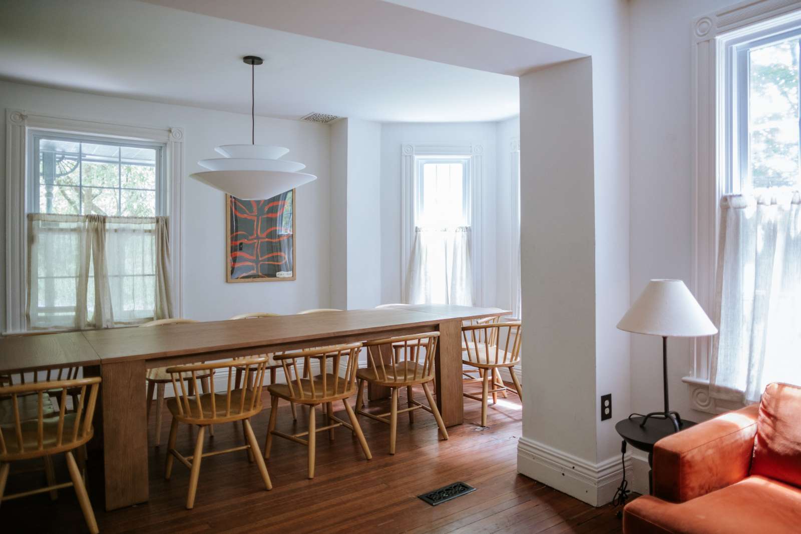 Spacious dining room featuring a long, light wooden dining table surrounded by numerous wooden spindle-back chairs. Large windows with sheer curtains and a modern pendant light fixture illuminate the room.