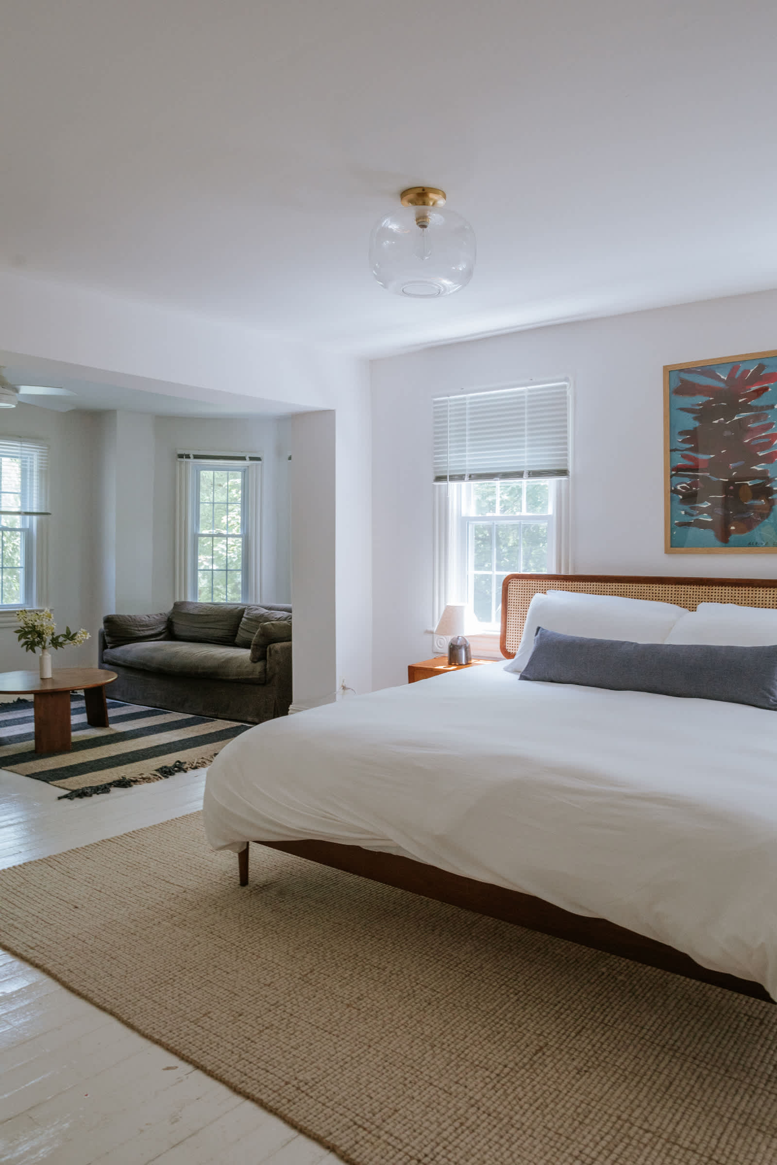 A spacious bedroom with white walls and light-painted hardwood floors, featuring a large bed with a cane headboard and crisp white bedding. In the background, a separate sitting area with a dark sofa and a round coffee table is visible through an archway.