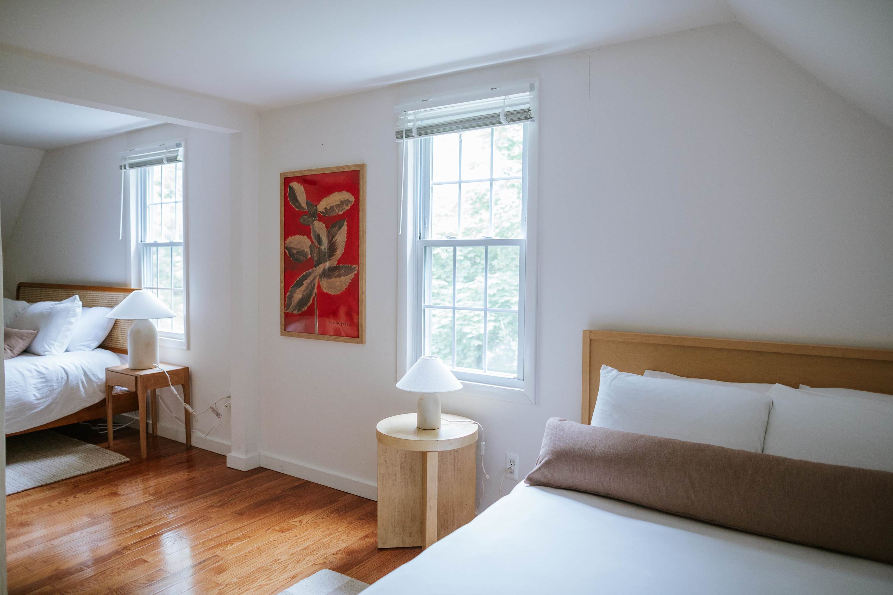 A bright room with white walls and hardwood floors features two distinct sleeping areas, each with a light wood bed frame, white bedding, a round side table, and a white table lamp, separated by a wall opening. A red botanical artwork hangs on the wall.