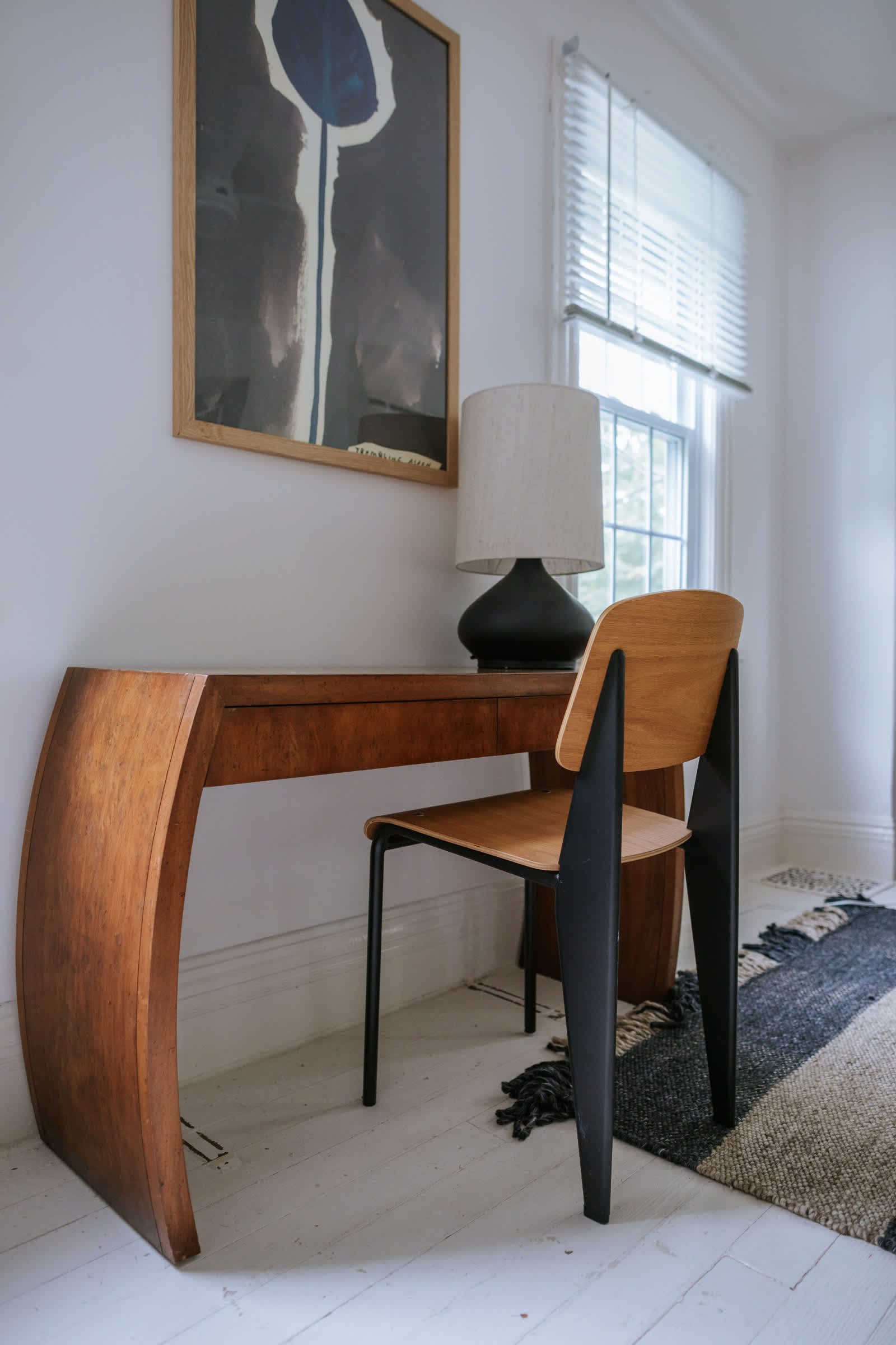 A stylish wooden desk with unique curved legs and a modern wooden chair with black accents, positioned against a white wall. A large framed abstract artwork hangs above the desk, and a table lamp sits next to a window with white blinds.