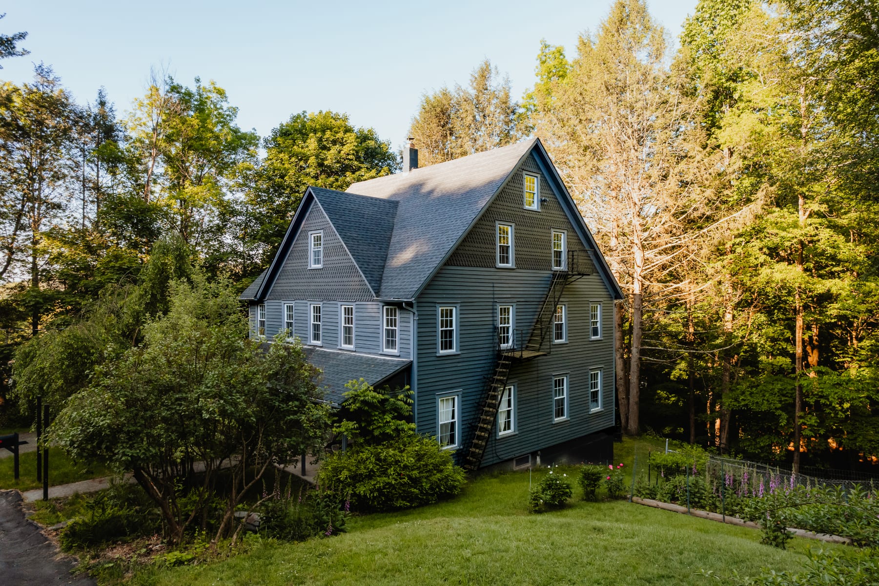 An aerial view of the large, dark blue farmhouse with multiple windows and a black metal fire escape on the side, surrounded by lush green trees and grass under a clear sky. The house has a gabled roof and shingle siding.