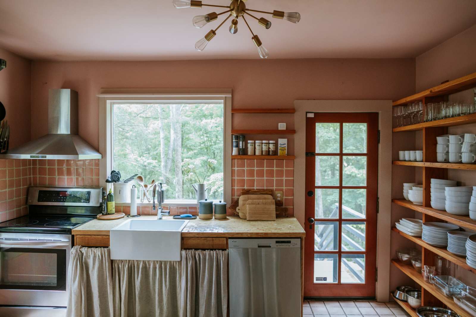 Rustic kitchen with a farmhouse sink, light wood countertops, and a terracotta-tiled backsplash. Open wooden shelving displays dishes, and a red door leads to an outdoor deck with a view of trees.