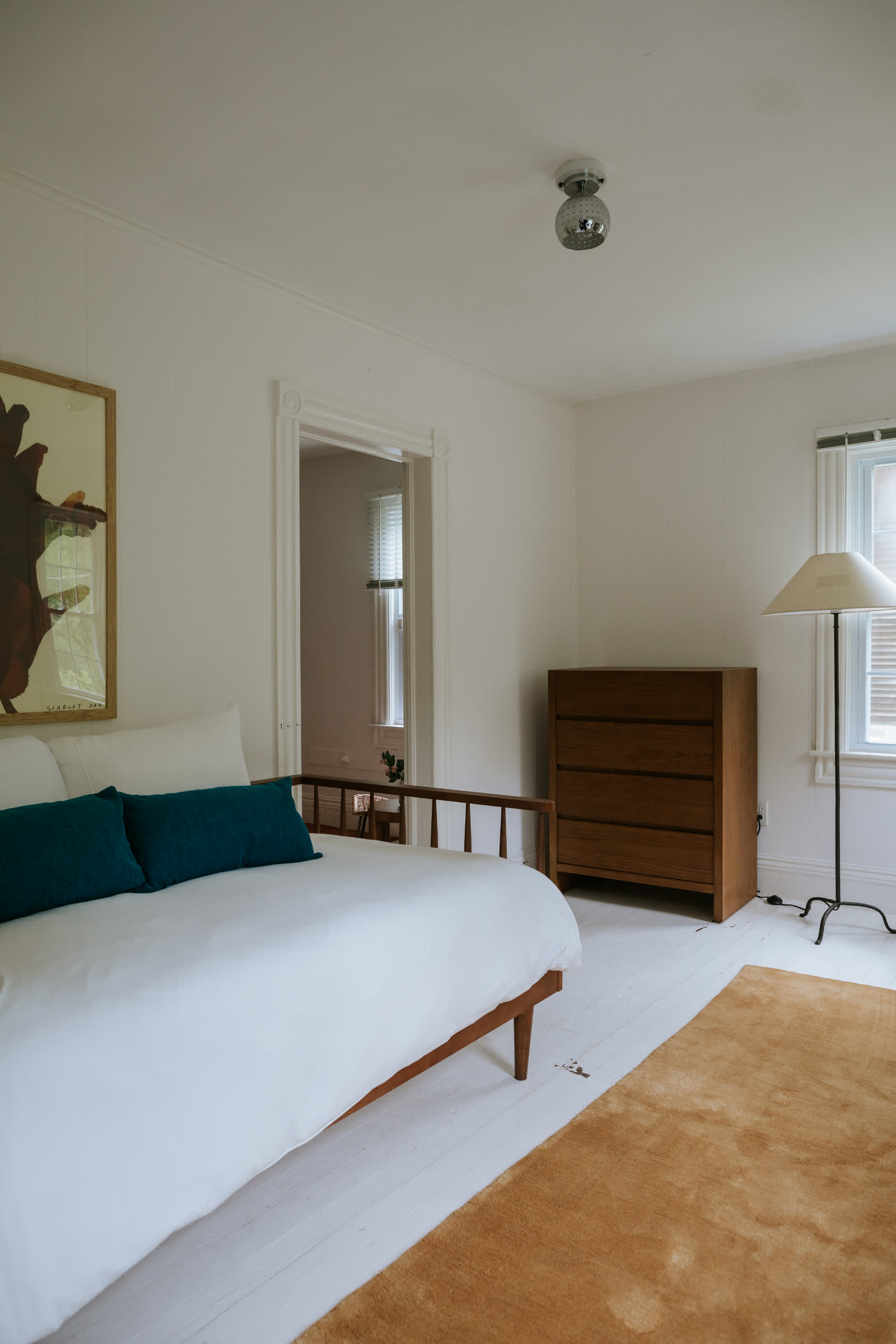 A bright bedroom features white paneled walls and painted white wood floors, a wooden spindle bed with white bedding and teal pillows, a matching wooden dresser, and a tall floor lamp beside a window.