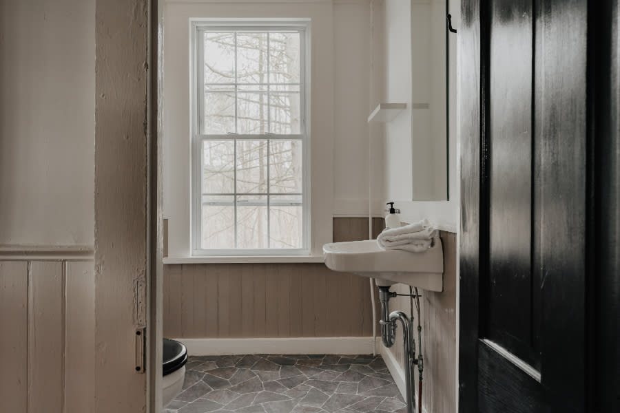 A bright bathroom featuring a white pedestal sink with exposed chrome plumbing, a toilet, and a large multi-pane window looking out onto bare trees. The floor is covered in grey geometric tiles, complemented by light brown wainscoting on the lower walls.