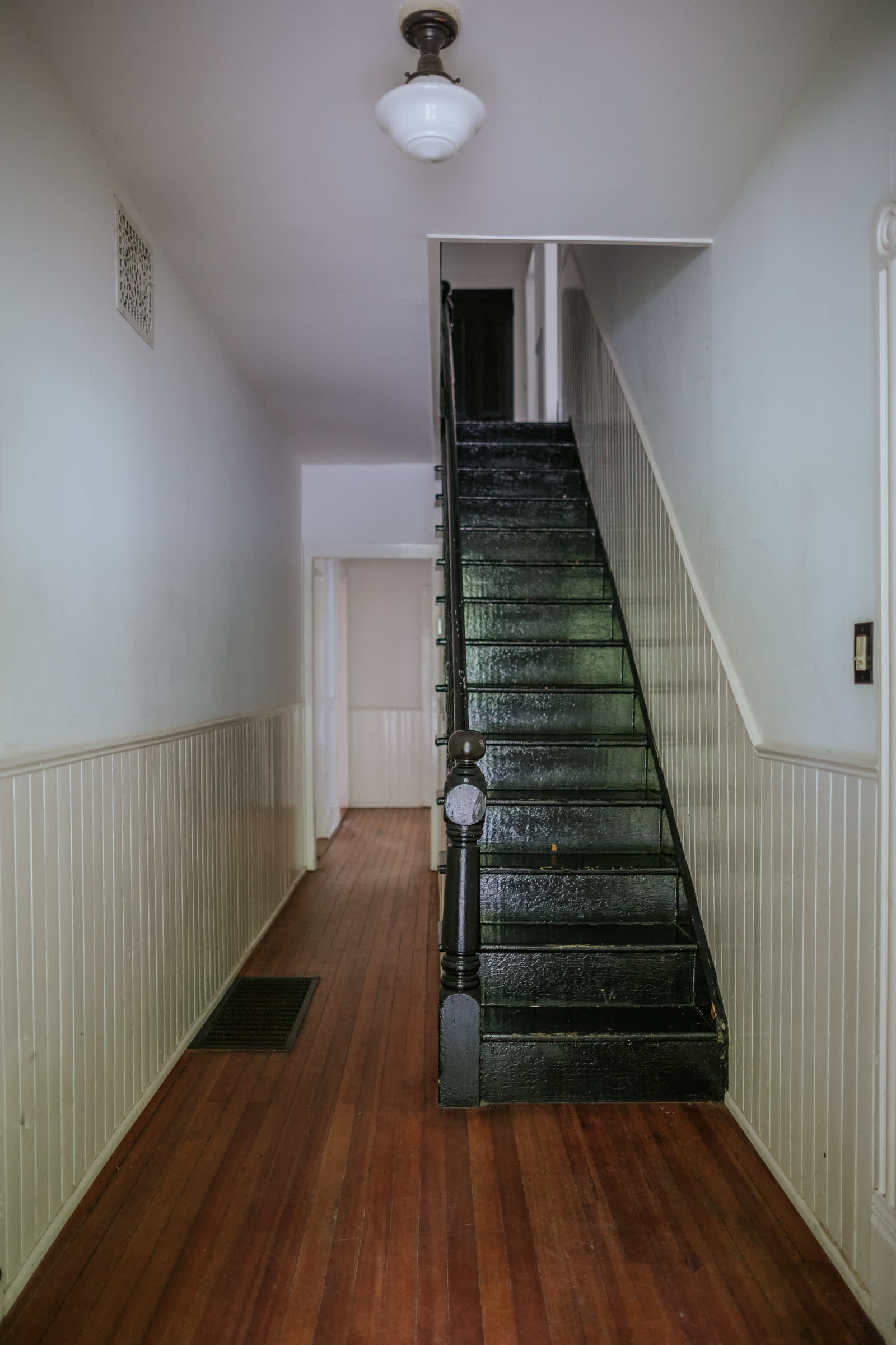 A long, narrow hallway with warm-toned hardwood floors and white wainscoting, leading to a steep, dark-painted staircase with a matching black banister. A vintage-style glass ceiling light fixture illuminates the space.