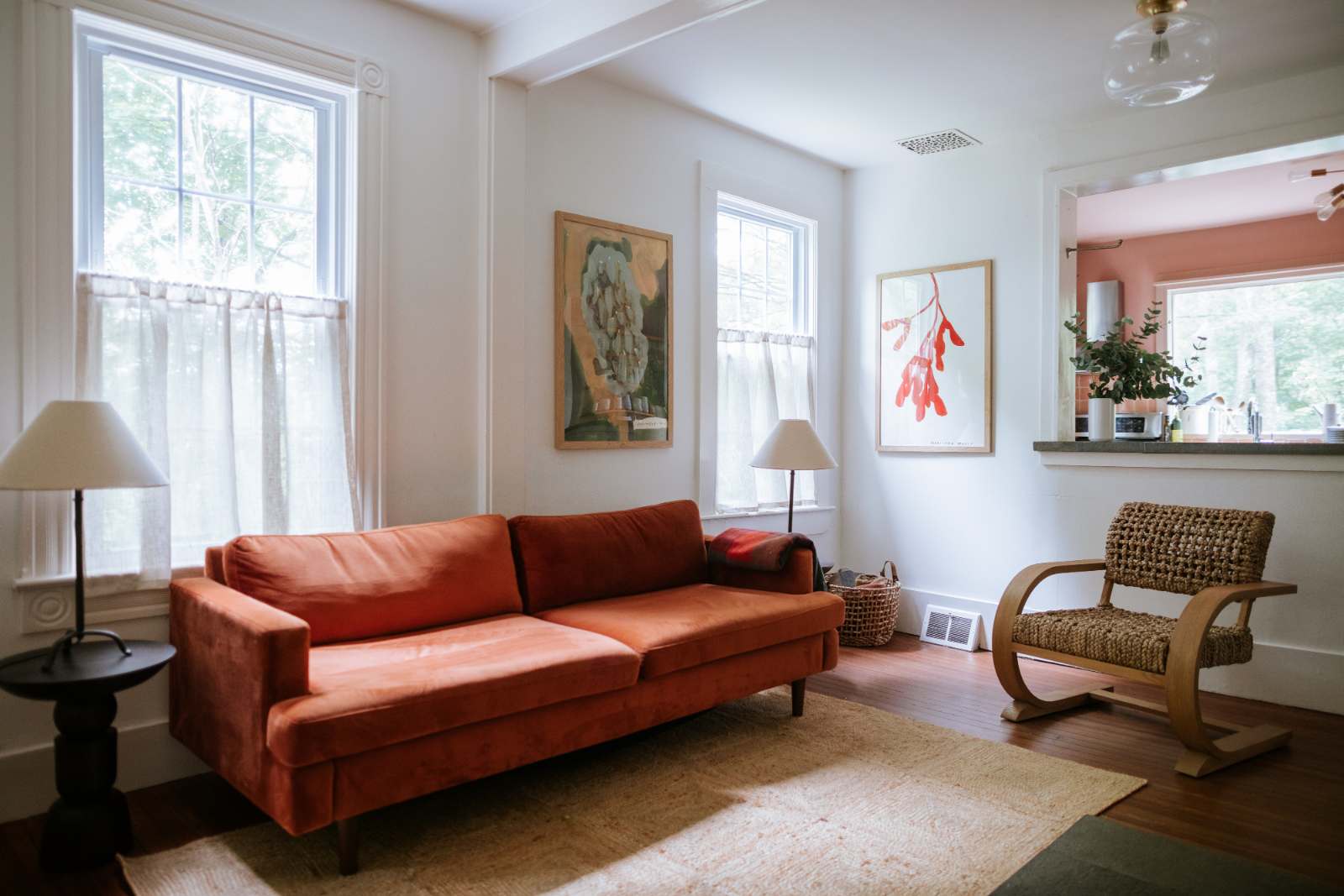 Cozy living room with a vibrant orange velvet sofa, two large windows with sheer curtains, and two framed art pieces on white walls. A small side table with a lamp and a woven chair complete the space.