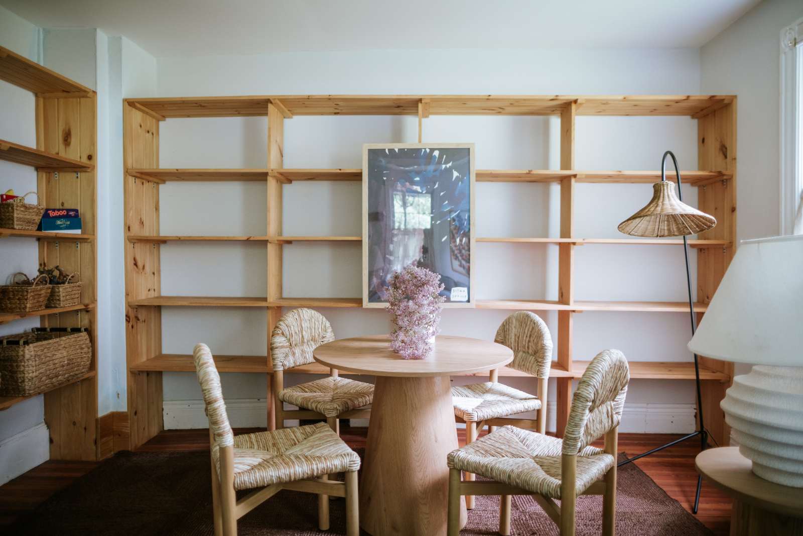 Bright room with extensive light wooden shelving units covering two walls, a round wooden table, and four chairs with woven seats. A framed picture hangs on the wall, and a vase with flowers is on the table.