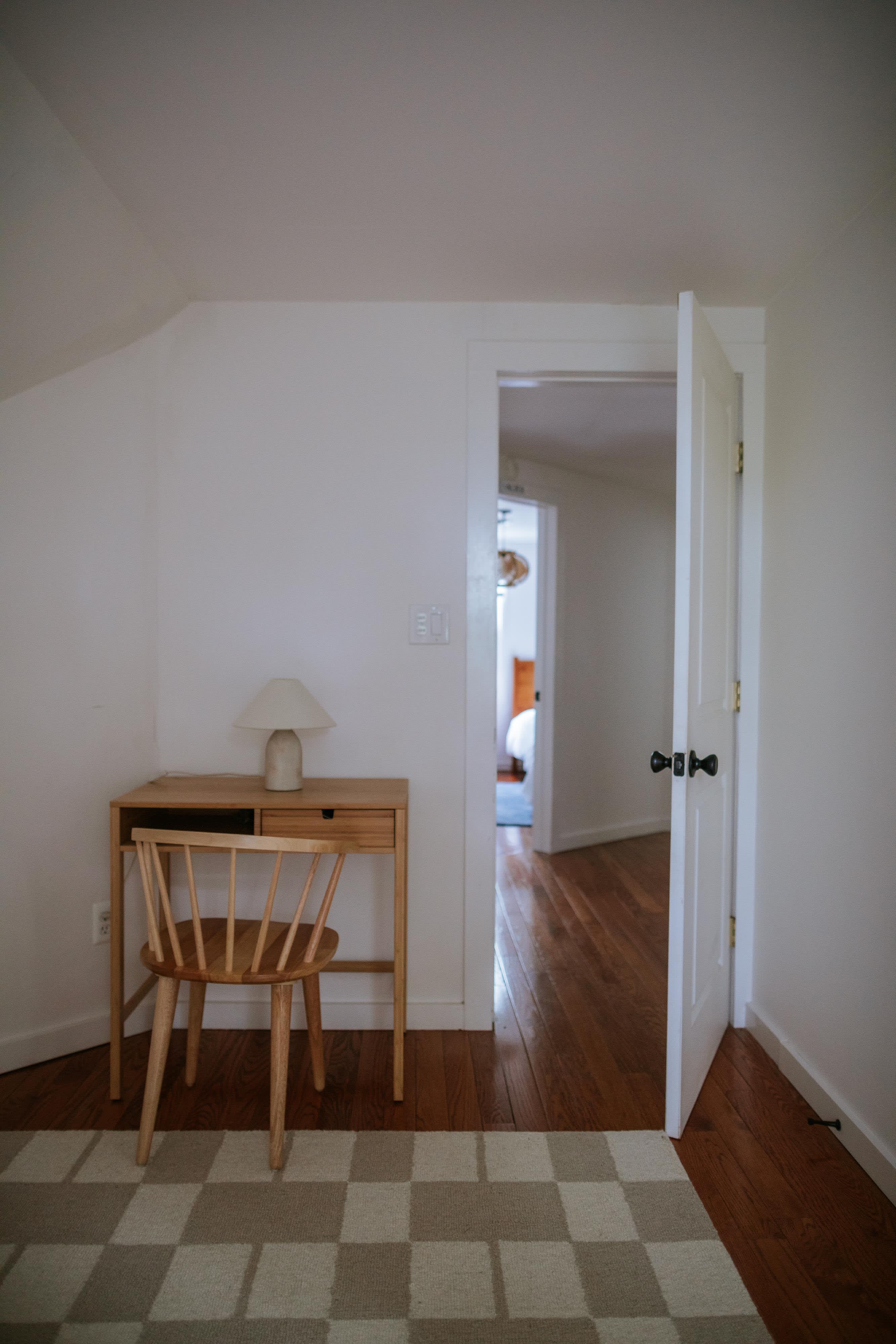 A small, bright nook with white walls and hardwood floors features a light wood desk and a matching wooden spindle chair, with a small white table lamp on the desk. An open doorway leads to another room.