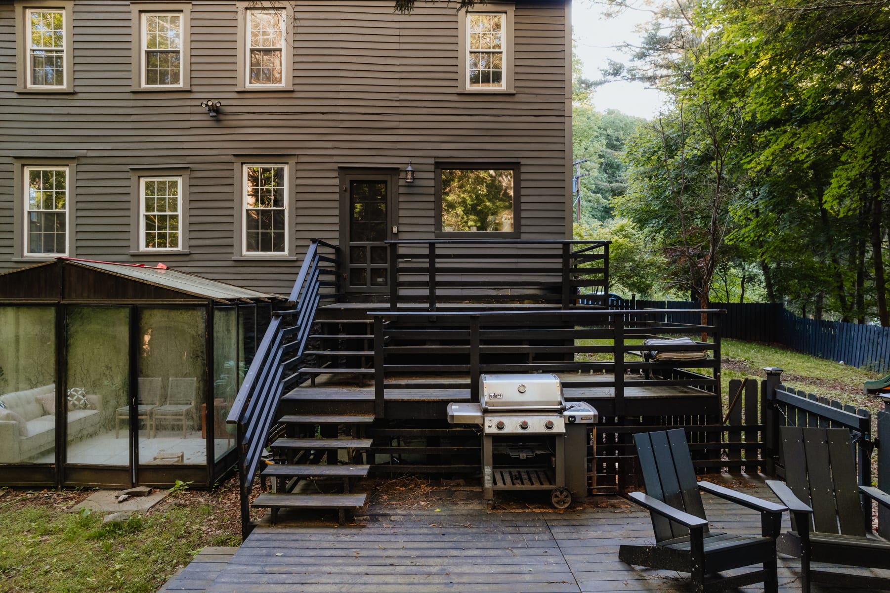 The back of a dark gray farmhouse features a multi-level wooden deck with black railings, a BBQ grill, and two black Adirondack chairs, with a glass greenhouse visible to the left.