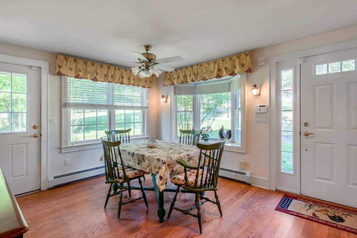 Inviting dining area with a wooden table and green chairs, surrounded by multiple windows with valances, and two white exterior doors, featuring hardwood floors and a ceiling fan.