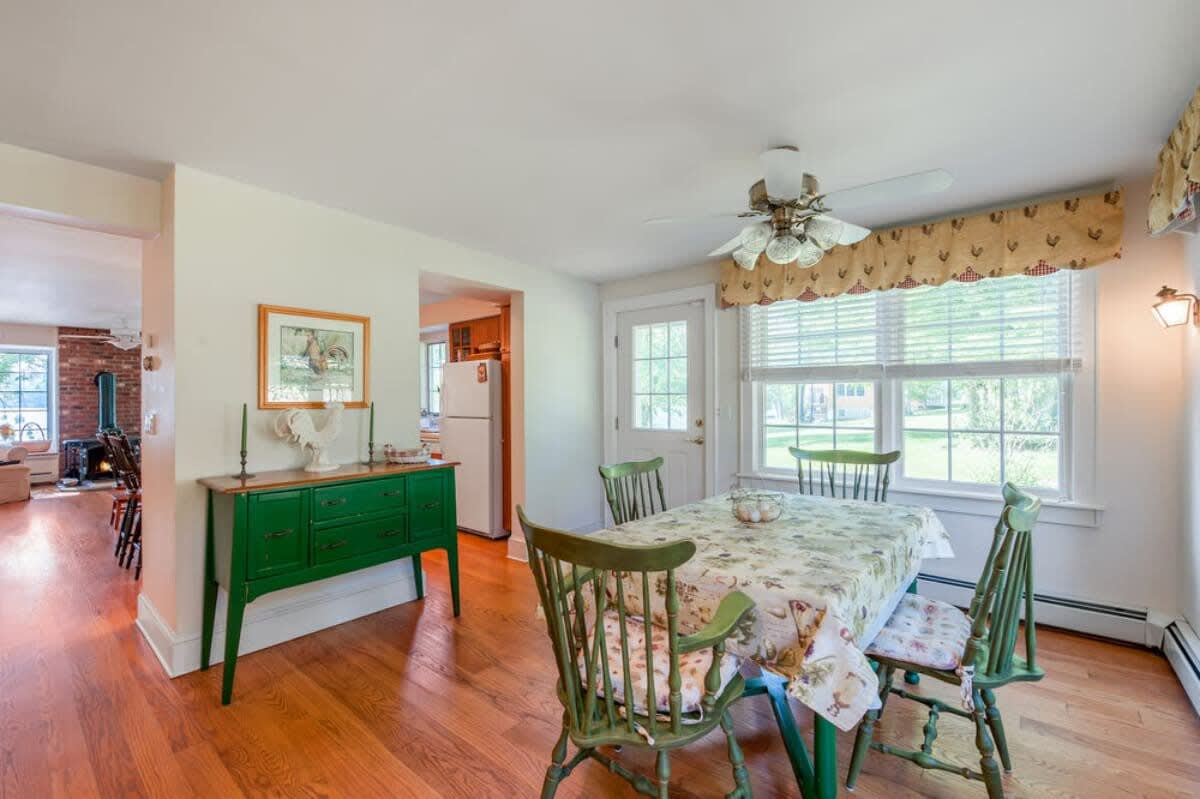 Charming dining area with a wooden table covered in a floral tablecloth, surrounded by green wooden chairs, and a vibrant green buffet cabinet, with hardwood floors and a view into the kitchen.