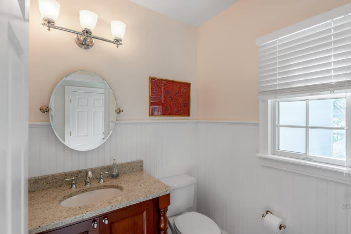 A compact bathroom with light peach walls above white beadboard wainscoting, a dark wood vanity with a granite countertop and oval sink, and a round mirror.