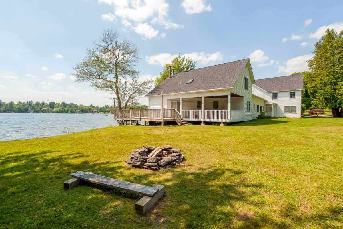 A white house with a dark roof and covered porch overlooks a lake, with a stone fire pit and a rustic wooden bench on a sprawling green lawn. Mature trees provide shade under a bright, partly cloudy sky.