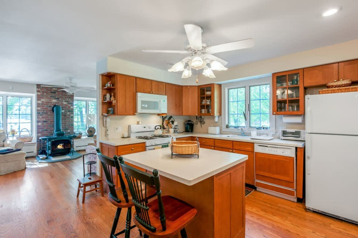 Bright kitchen with light wood cabinetry, white countertops, a central island with two bar stools, and stainless steel appliances, opening into a living area with a brick fireplace.