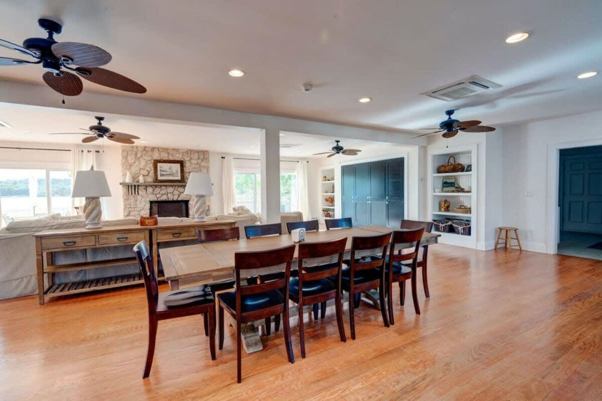 Spacious dining area with a long wooden table and dark chairs, open to the living room with a stone fireplace, featuring hardwood floors and multiple ceiling fans.