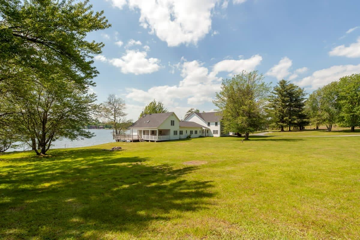 A sprawling white house with a covered porch and deck overlooks a calm lake, surrounded by a vast, sunlit green lawn and scattered mature trees under a partly cloudy blue sky. The property extends to the lake's edge, highlighting its waterfront location.