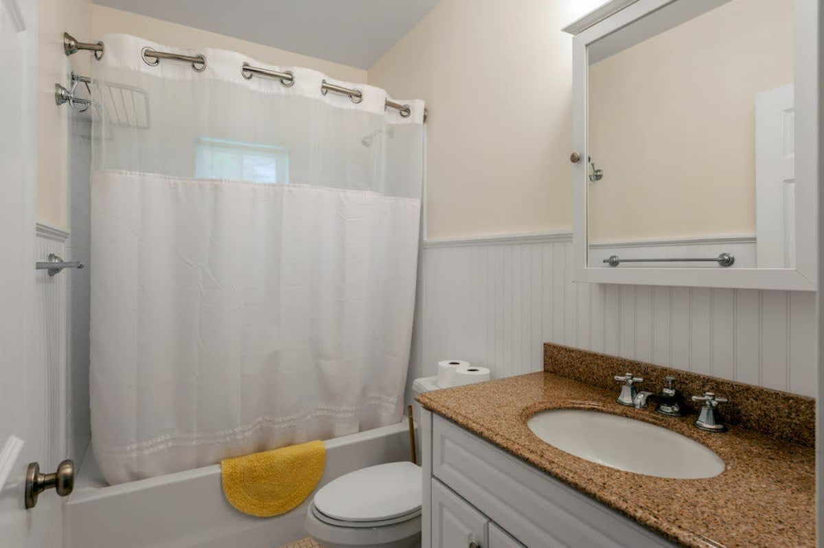 A bright bathroom featuring a white bathtub with a clear and white shower curtain, a light-colored vanity with a granite countertop and a single sink, and white beadboard wainscoting.