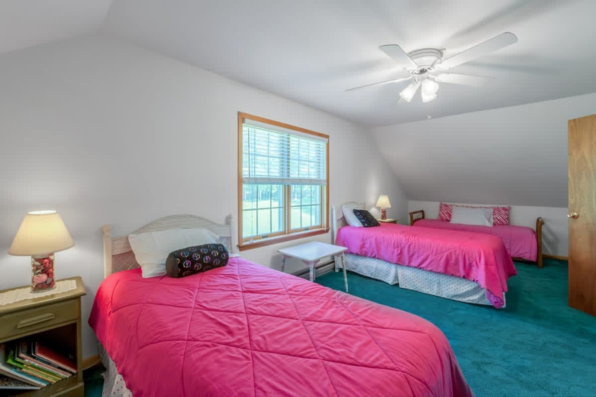 Bright bedroom with two twin beds featuring vibrant pink comforters, a large window with blinds, and a sloped ceiling, set on a teal carpeted floor.
