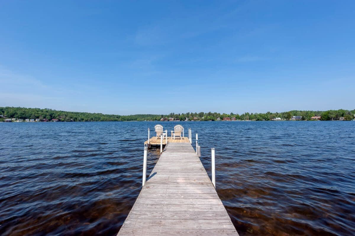 A long wooden dock extends straight into a vast, calm lake under a clear blue sky, with two white Adirondack chairs positioned at the end, offering a perfect spot for enjoying the expansive water views. Distant tree-lined shores frame the horizon.
